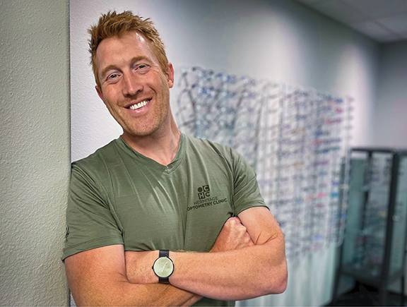 Dr. Sleichter leaning with arms crossed against corner in front of wall of glasses frames