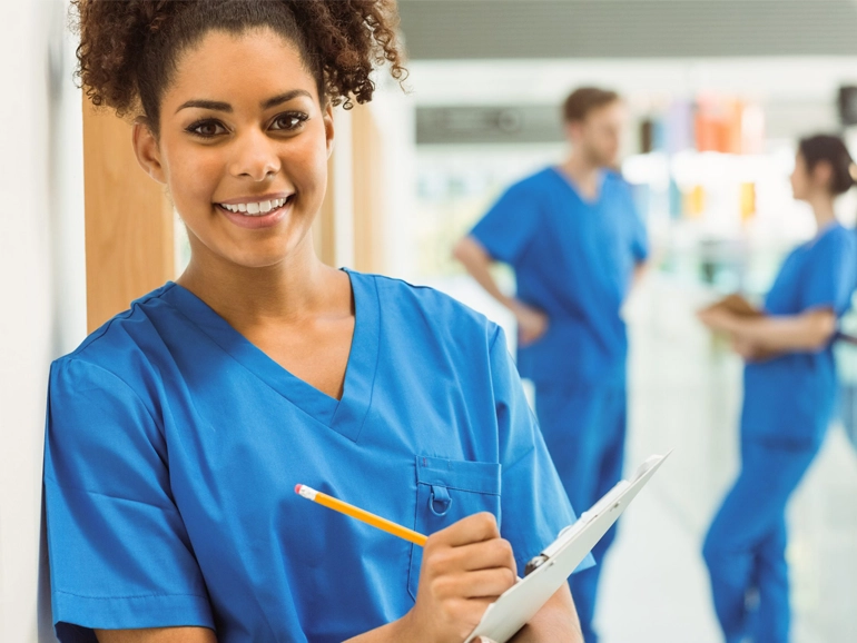 Healthcare student wearing scrubs and standing in hallway with pencil and clipboard
